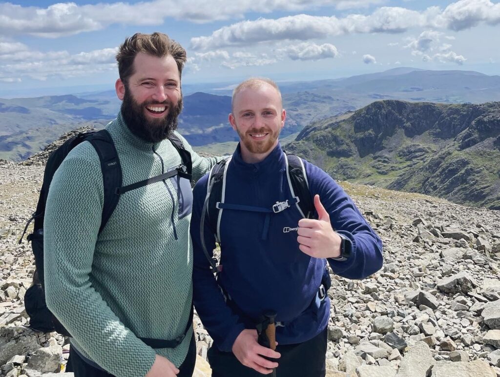 Two men standing on top of a hill in walking gear, smiling at the camera