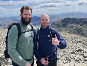 Two men standing on top of a hill in walking gear, smiling at the camera