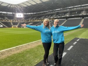 Two women dressed in blue hoodies standing at the edge of a football pitch smiling.