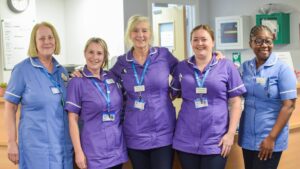 Five nurses wearing purple and blue uniform smile at the camera.