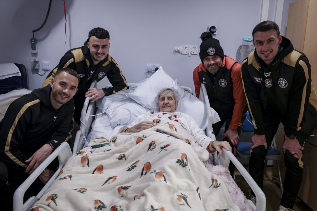 Football players stand around a hospice bed smiling and the camera. A lady in bed is also smiling.