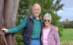 Older man and woman smiling for camera outside by a metal structure